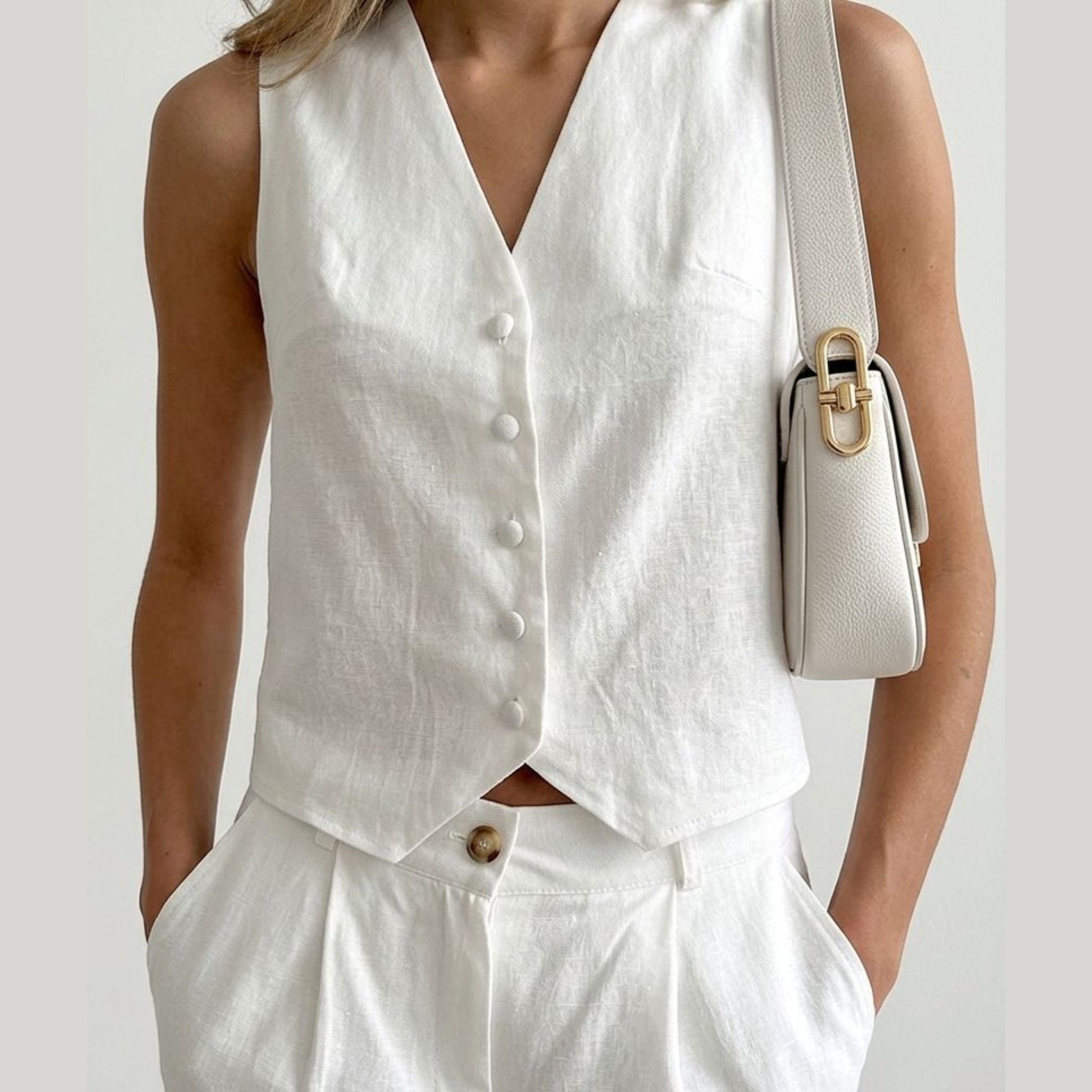 Person wearing a white sleeveless top and pants with a beige handbag on a plain background